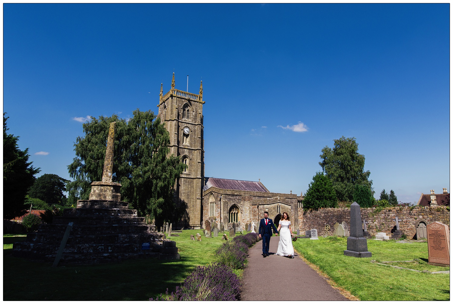 Bride and groom holding hands and walking up the path from St Andrew's Church, Chew Magna. The sky is very blue.