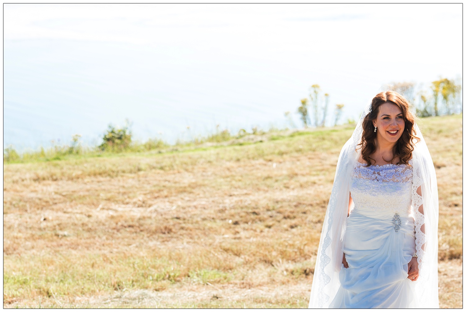 The bride by Chew Valley Lake in the sunshine. She's hitching up her dress as she walks on the grass.