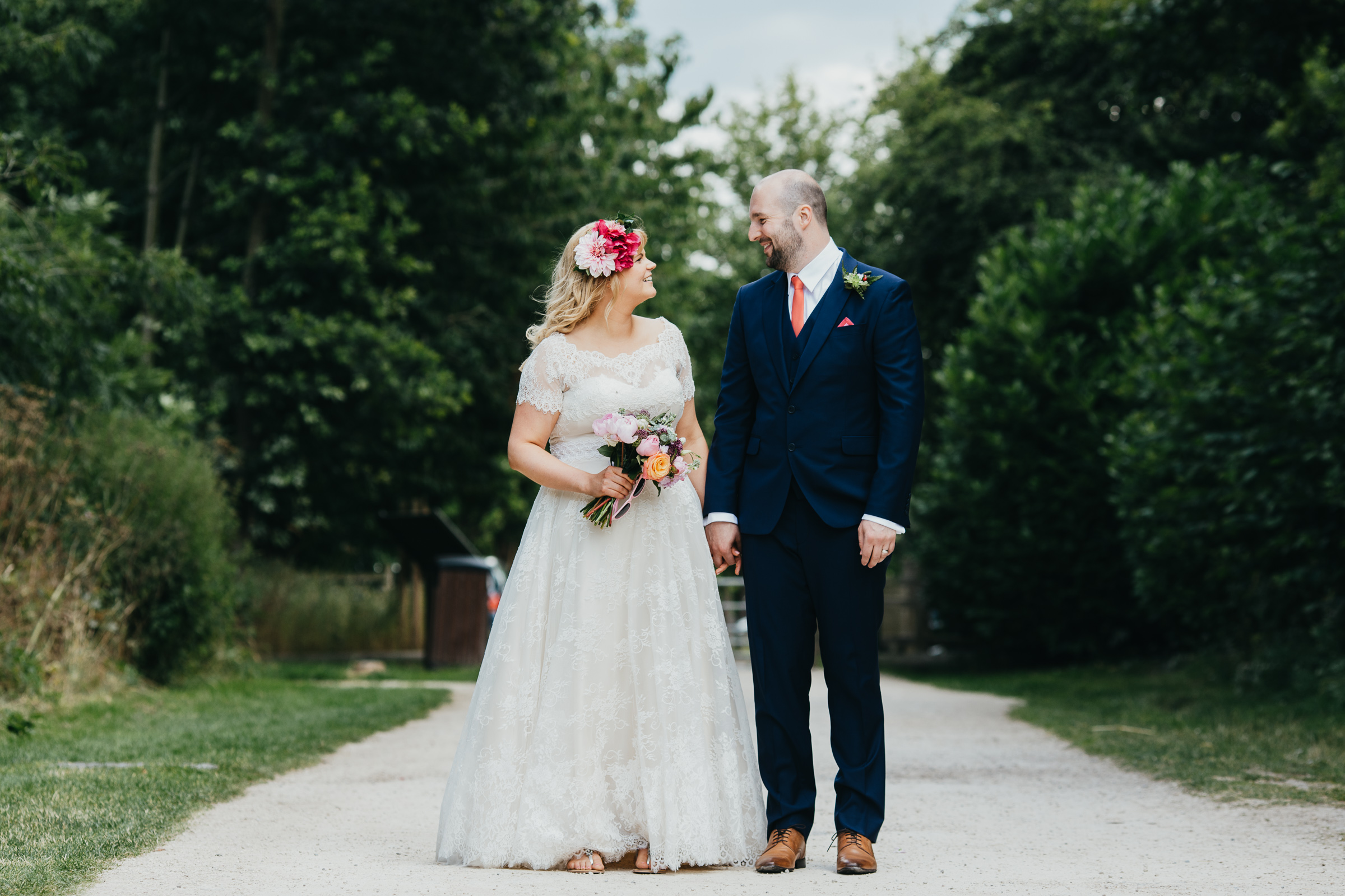A woman in a wedding dress and flowers in her hair is holding hands with a man. He is wearing a blue suit with a coral coloured tie. They just got married. they stand in the path of Rushcliffe Country Park.