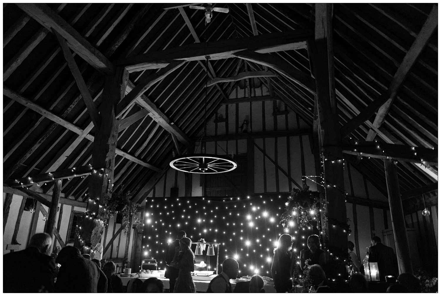 The view of Pledgdon Barn at night. There is a wall of lights behind a buffet table. People are walking around and chatting.
