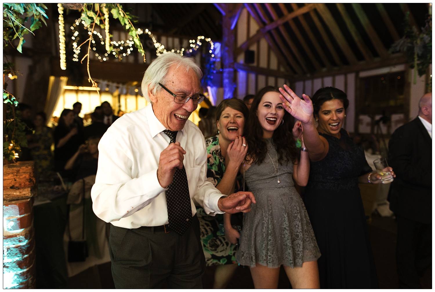 An elderly man is dancing at a wedding reception with three women. They are in Pledgdon Barn and there are fairy lights.