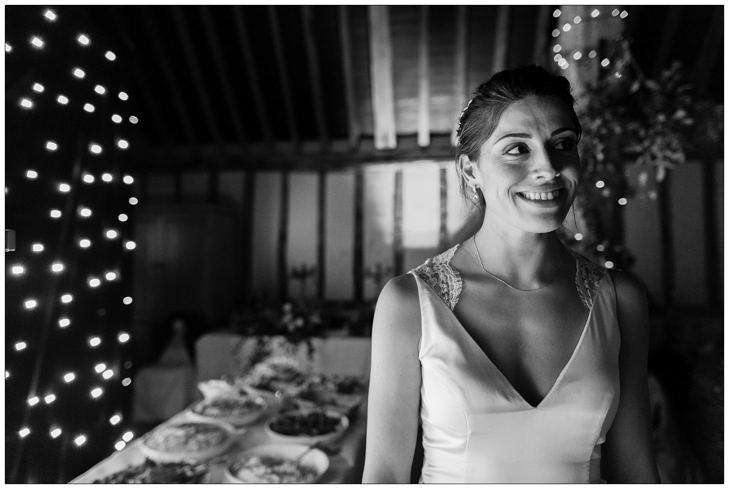A bride smiling by the buffet table and a wall of little lights. At Pledgdon Barn wedding.