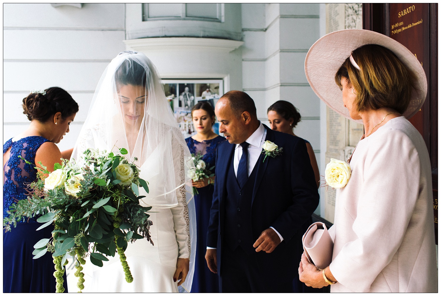 Bride behind her veil is looking down, her parents and bridesmaids are with her. She's about to marry in St. Peter's Italian Church in Clerkenwell.