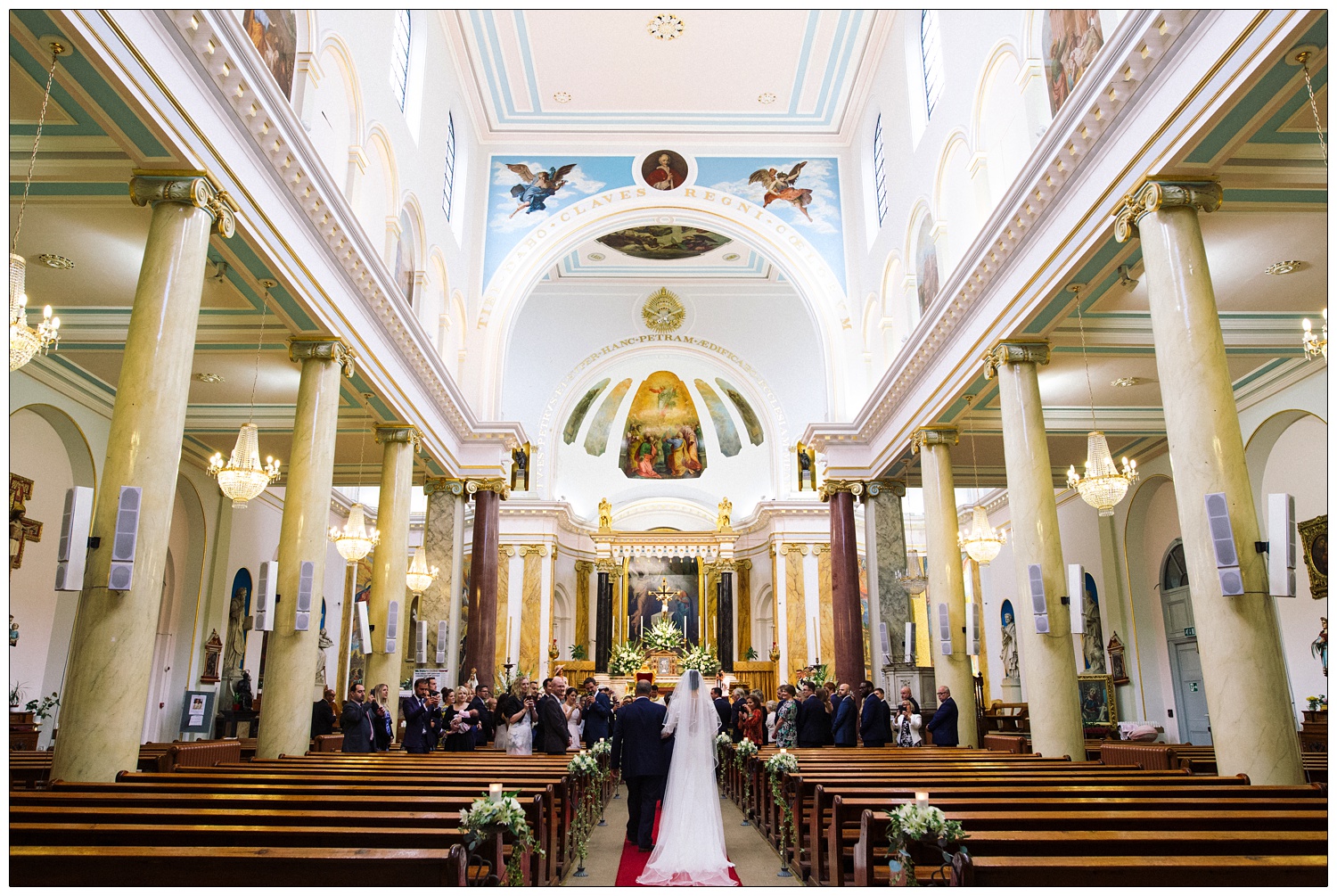 The bride being walking up the aisle with her father. The view from behind as the veil trails on the red carpet. St. Peter's Italian Church in Clerkenwell.