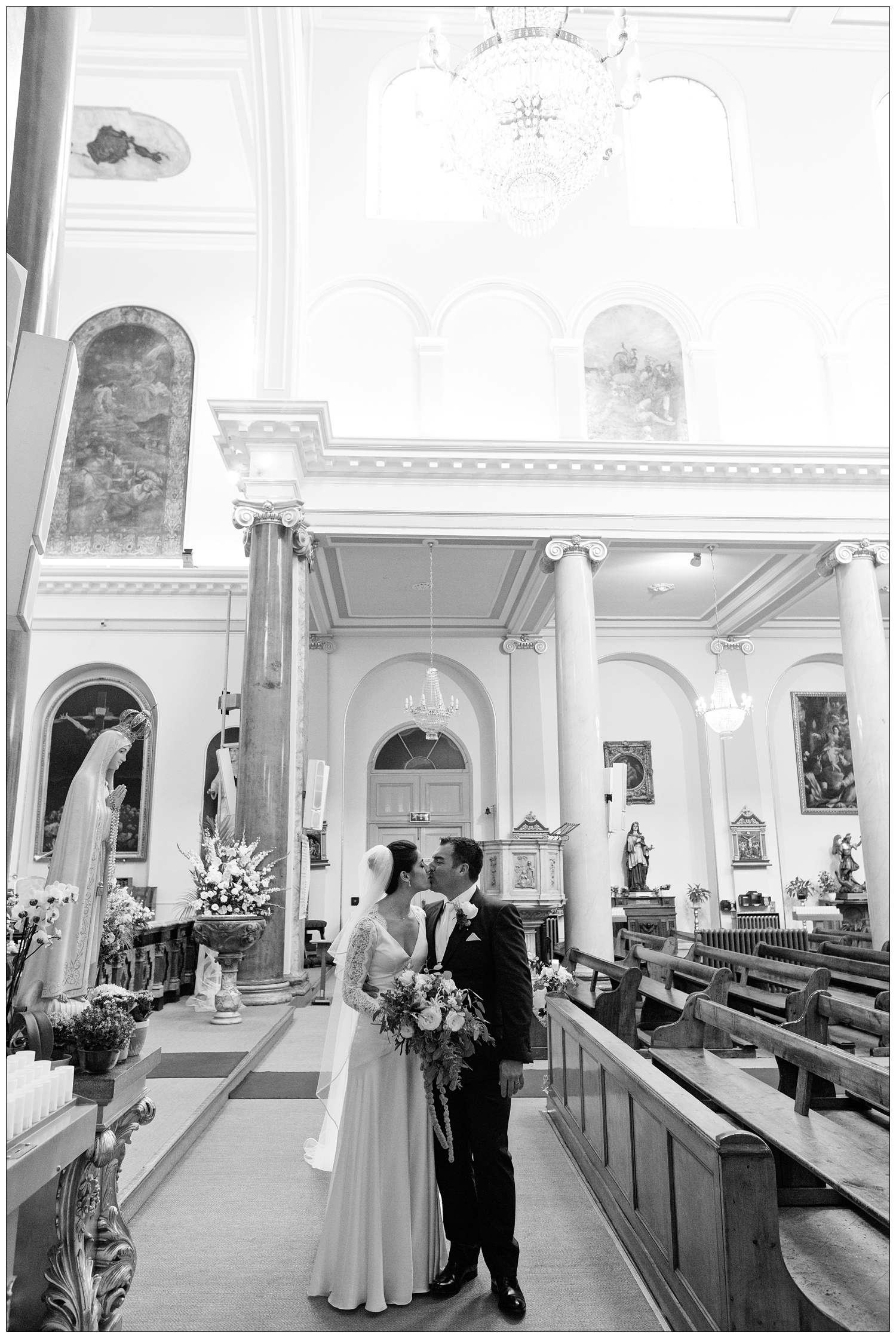 Bride in a long white wedding dress and veil kisses her husband in the St. Peter's Italian Church in Clerkenwell.