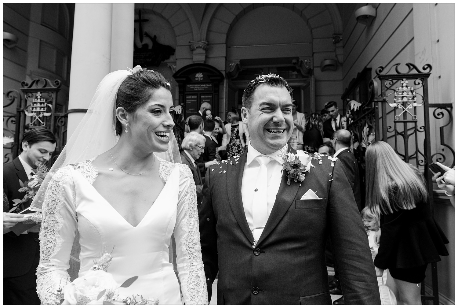 Woman in a wedding dress and veil, with a man in a suit. They are covered in confetti outside St. Peter's Italian Church in Clerkenwell.