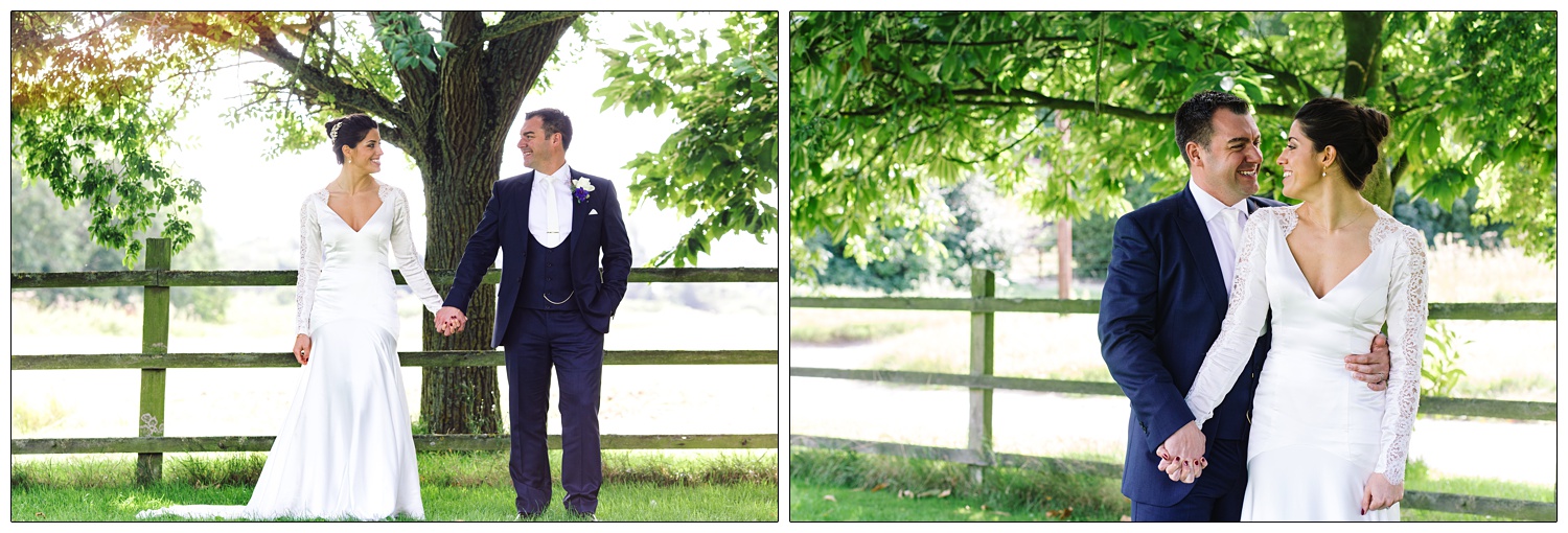 A newly married couple holding hands under a tree. She is wearing an elegant long satin silk dress with lace sleeves. He is wearing a blue three piece suit and a white tie.