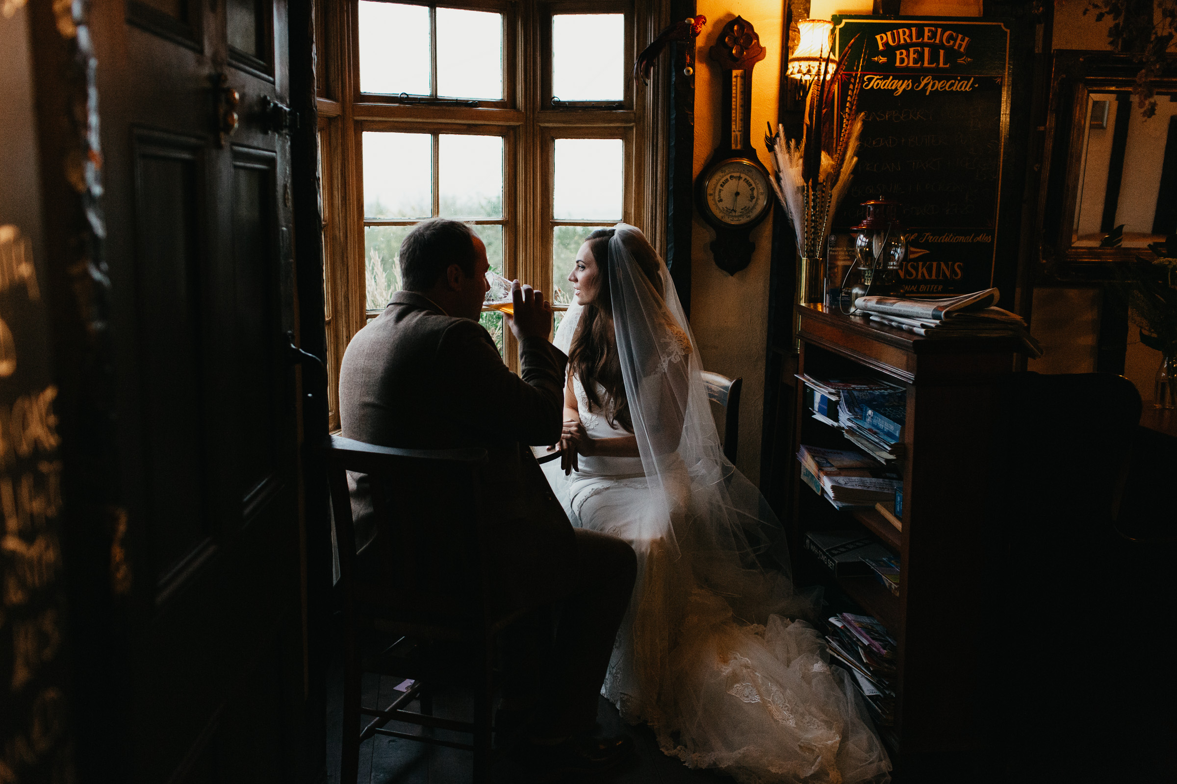 A bride is sat in the window of the Purleigh Bell in Essex looking out of the window. She is wearing a Galia Lahav dress. The groom is sat opposite his new wife and is drinking a pint.