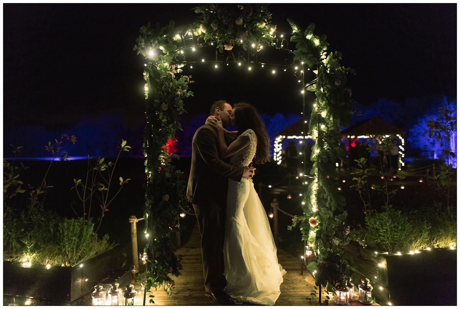 Under an arch that says Honeywood Farm a bride and groom kiss. The trees in the background are lit up blue.
