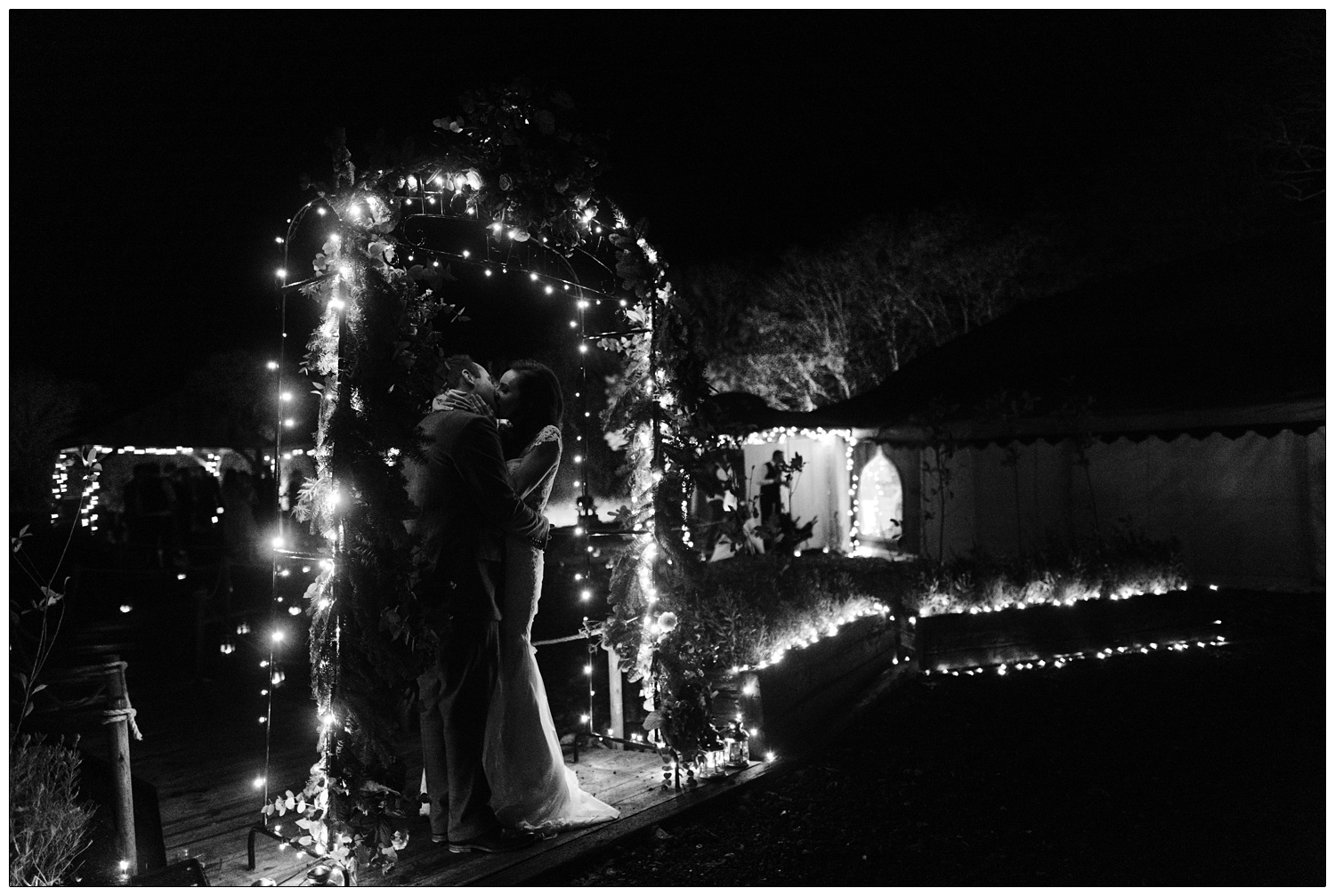 Bride and groom kiss under an arch with the words Honeywood farm at the top. There are fairy lights. The wedding orangery is in the background and it's night time.