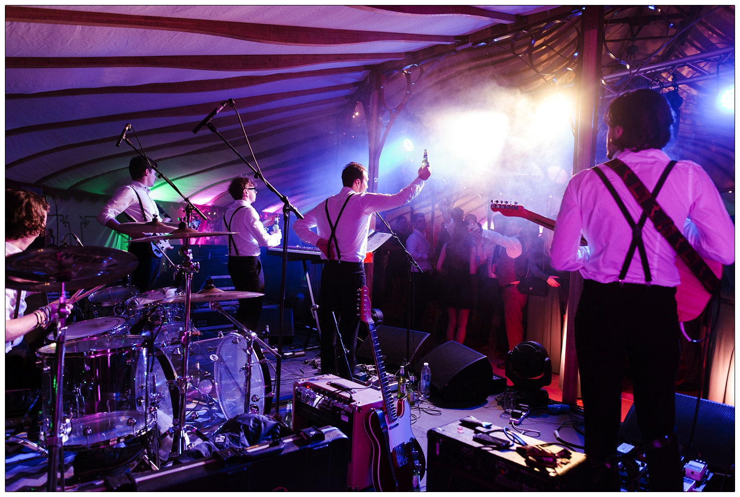 The view of Winston and The Lads wedding band from behind performing in an orangery ent. The singer Nick is raising a bottle of beer to the wedding crowd. The lights are mostly magenta.
