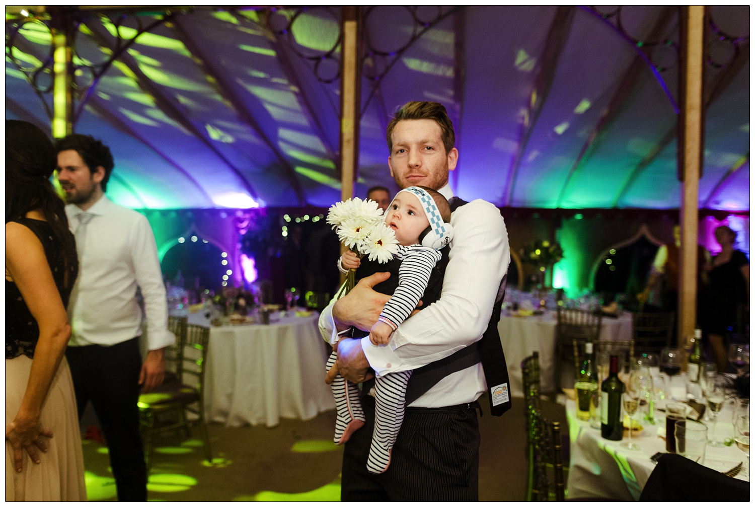 A man with his baby in a carrier. The baby is wearing ear defenders and is holding white flowers. They are at a wedding reception.