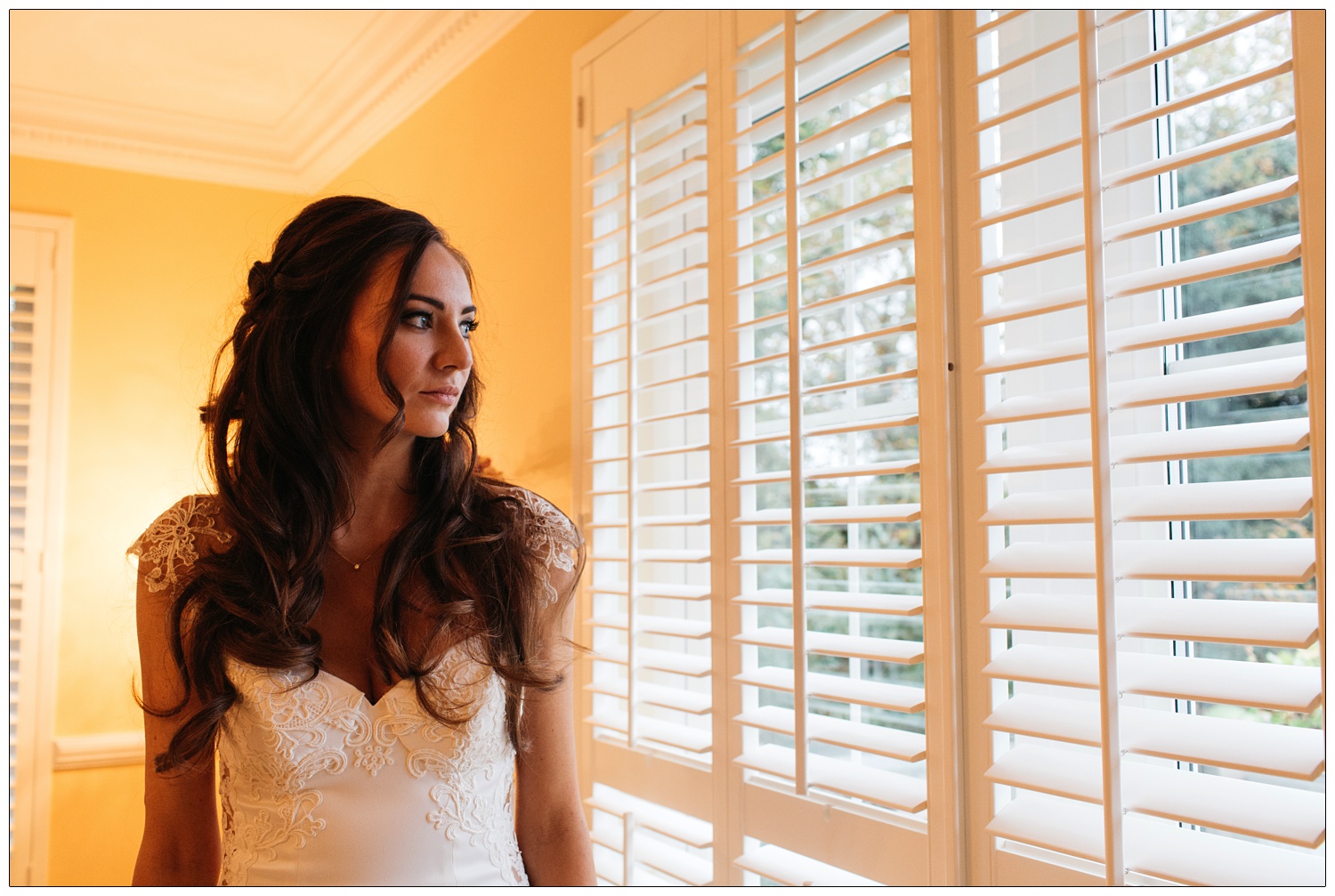 Bride with long dark curled hair, wearing Galia Lahav from Browns bride, looks out of the bedroom shutters.