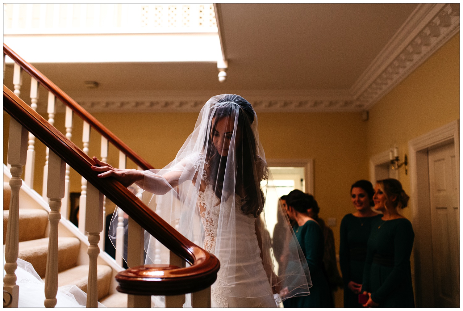 A bride in a Galia Lahav dress and veil walks down the stairs, her hand is on the bannister.