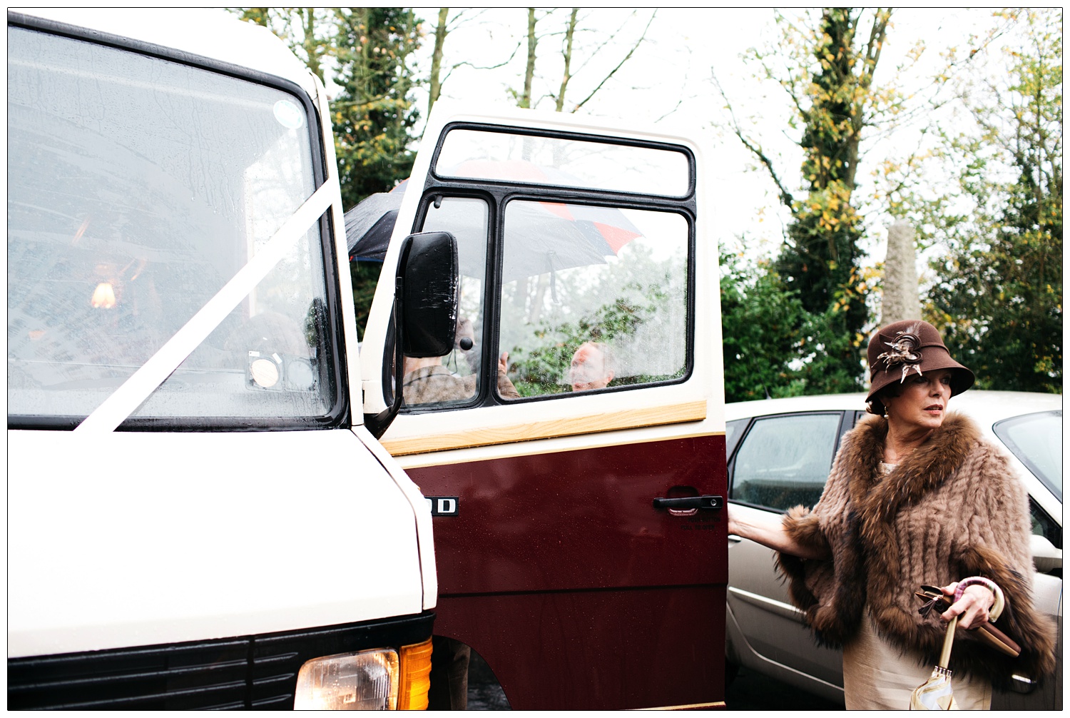 Mother of the groom in a brown furry shawl and hat is outside in the rain. About to get aboard a coach.