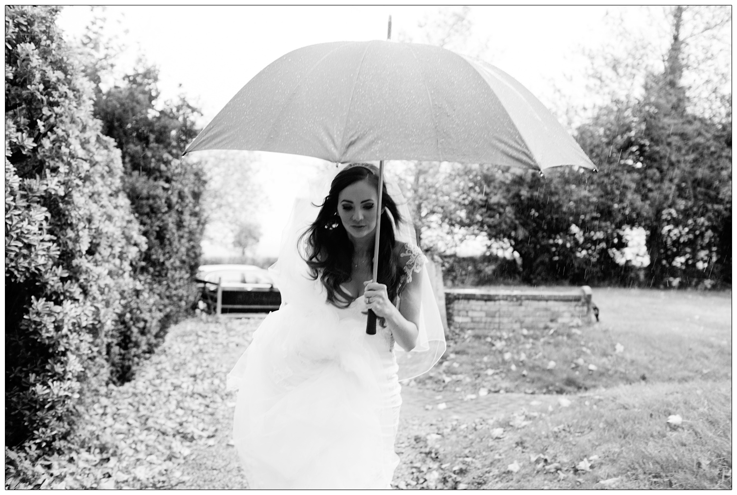 A bride in a white wedding dress and holding an umbrella. It's a windy and wet November day. She's walking up the path to the All Saints Church in Purleigh for her wedding ceremony.