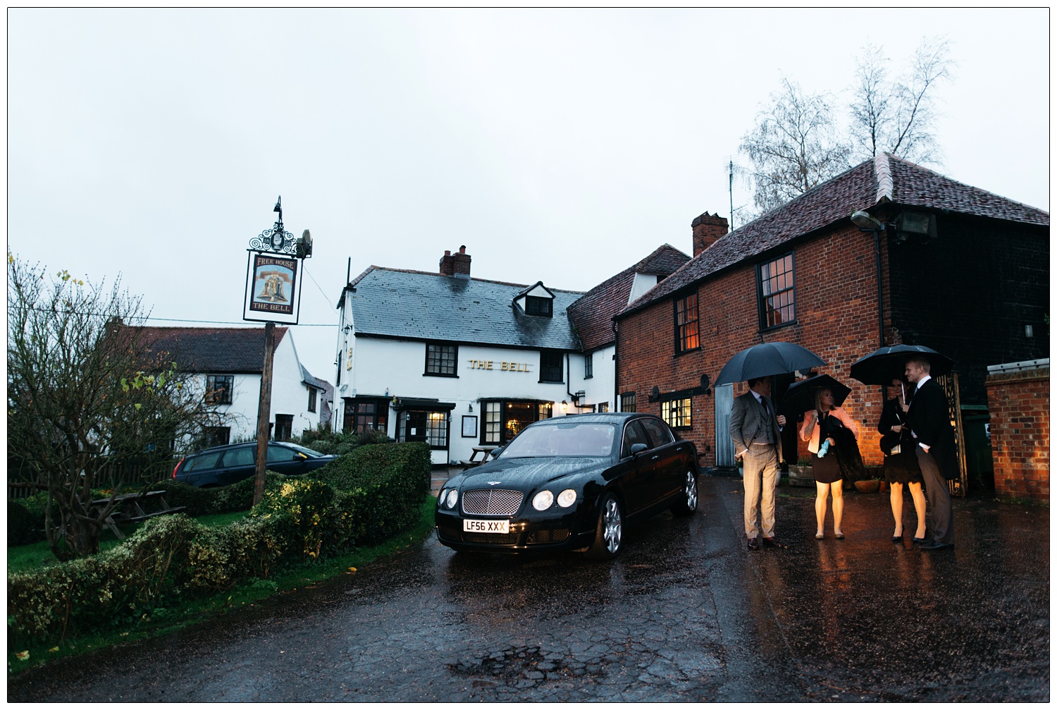 People are stood outside under umbrellas. There is a Bentley car. They are outside the Purleigh Bell.