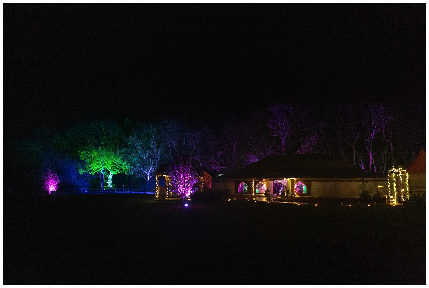 The orangery style tent for a wedding is in front of some trees. They are lit up in coloured lights. Magenta, blue, green and red. The path to the tent is lit up. It's is night time.