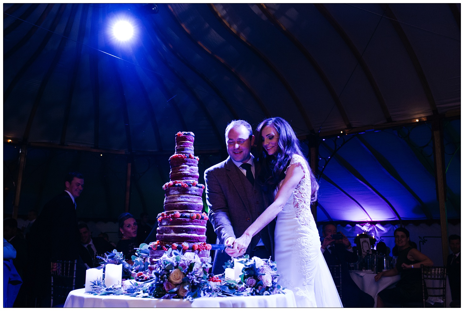The bride and groom cut their very tall wedding cake, under a blue spotlight.