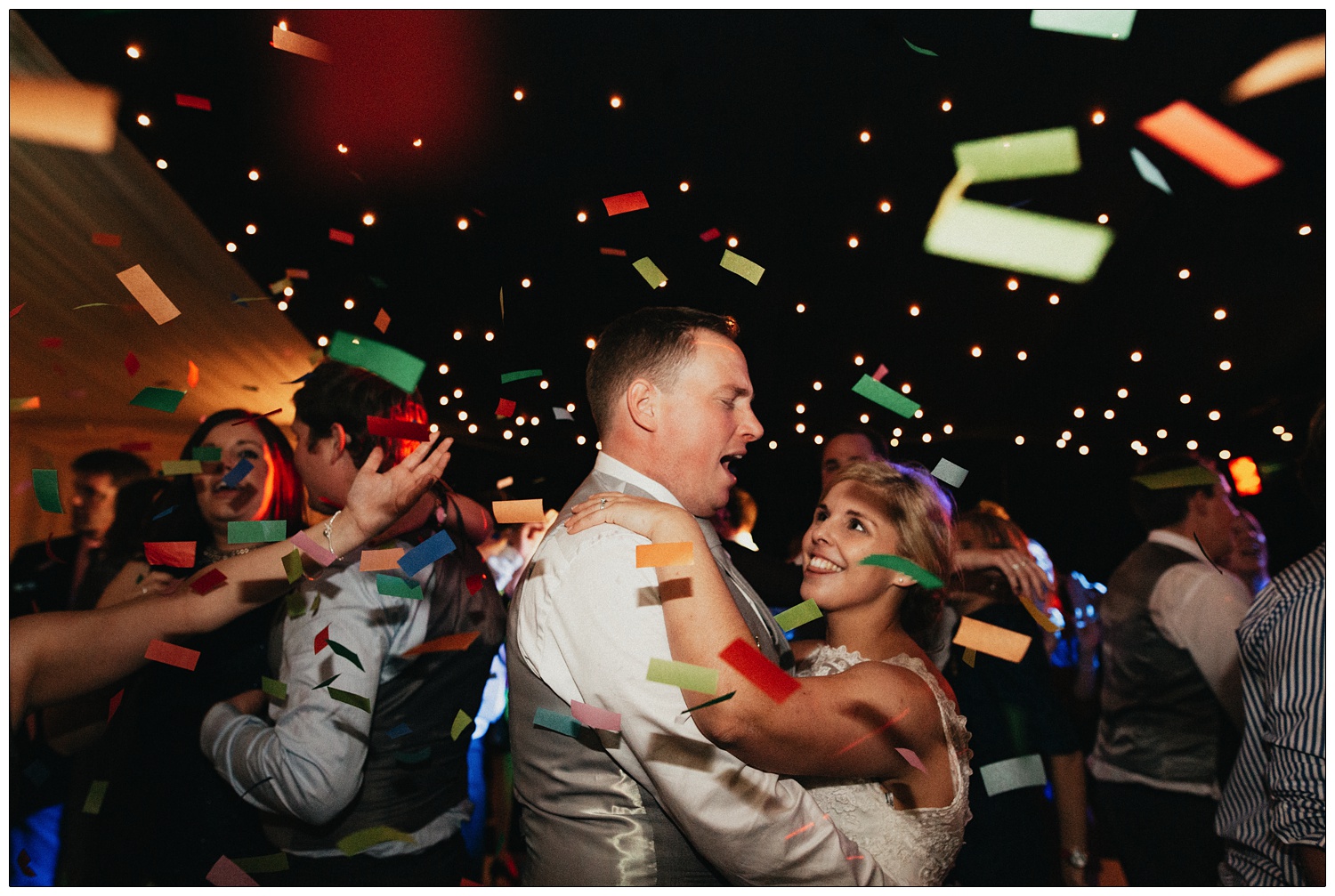 A confetti cannon goes off on a dance floor and showers the bride and groom in multi-coloured confetti. Wedding guests dance around them.