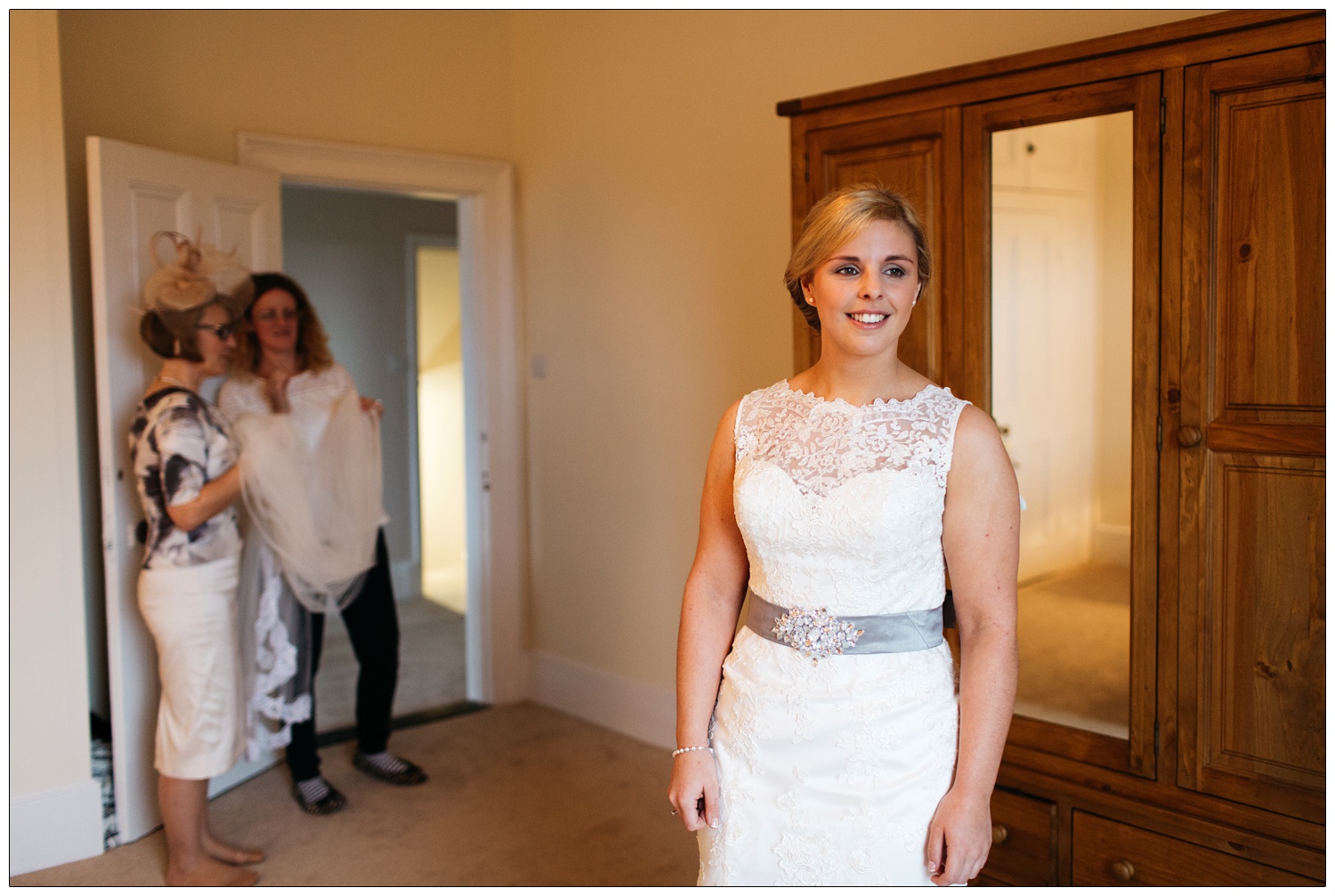 A bride stands in a bedroom in her dress. In the background two women ar holding the veil.