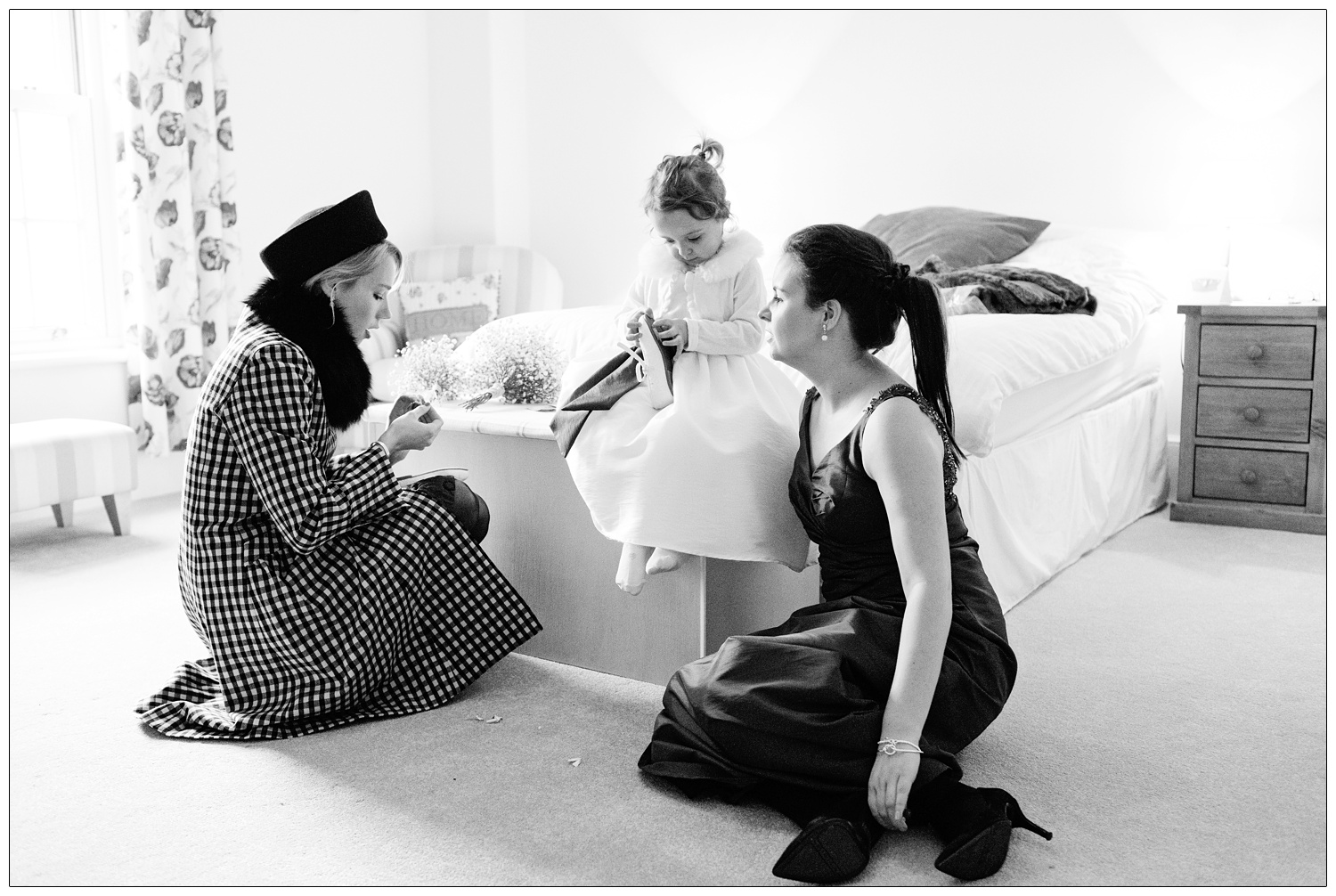 A flower girls is sat on a bed and picking the label from a pair of shoes. Two women are sat on the carpet.