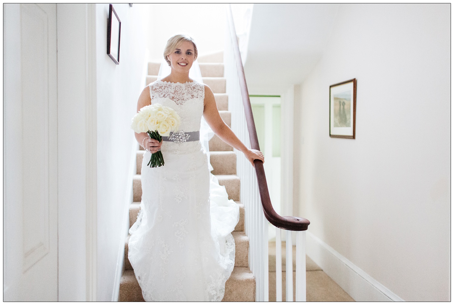 A bride in her lace dress carrying white roses is on the stairs at home.