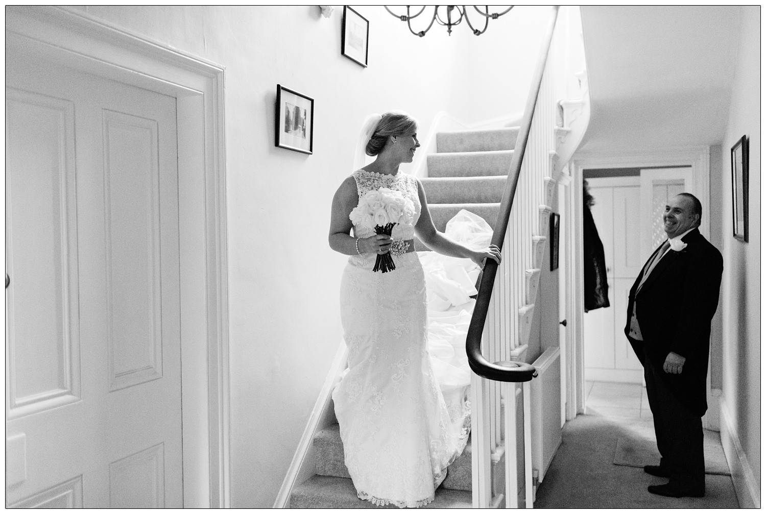 A woman in her wedding dress carrying flowers walks down the stairs of the family home and smiles at her dad.