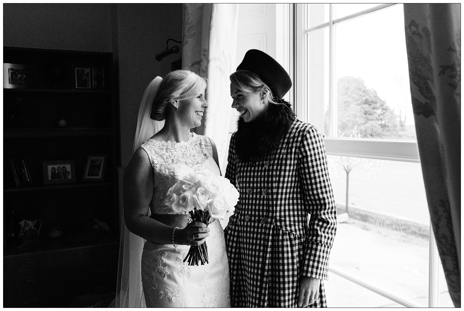 Bride and her friend stand in the window at home and smile at each other.