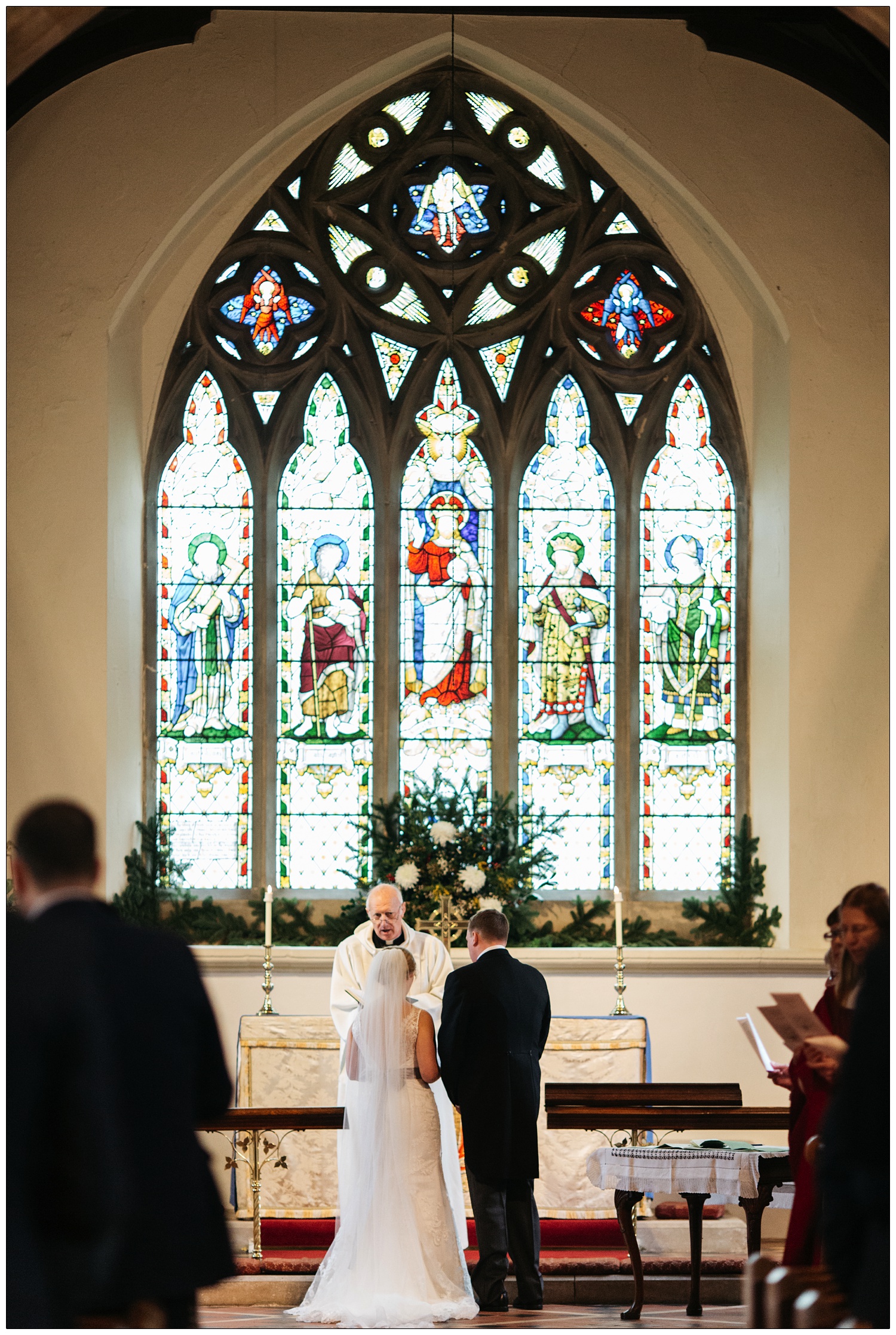 The bride and groom stand in front of the stained glass window and vicar at the St Thomas' Church in Bradwell-on-Sea
