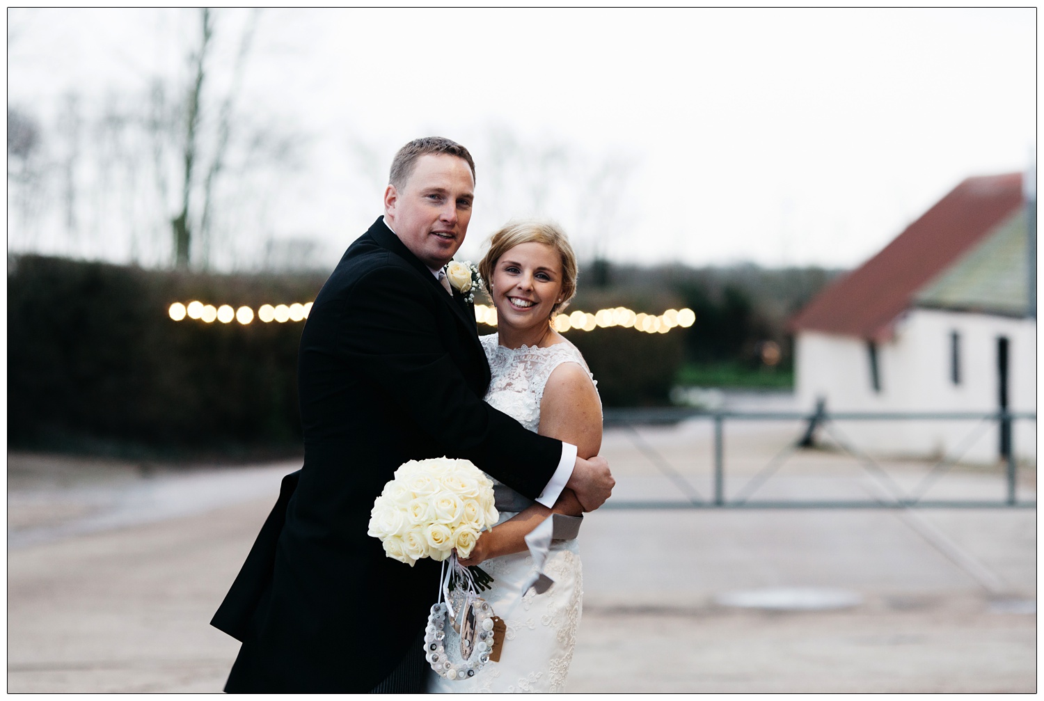 Groom hugs his bride on their chilly wedding day in January. There is a string of lights behind them on the farm.