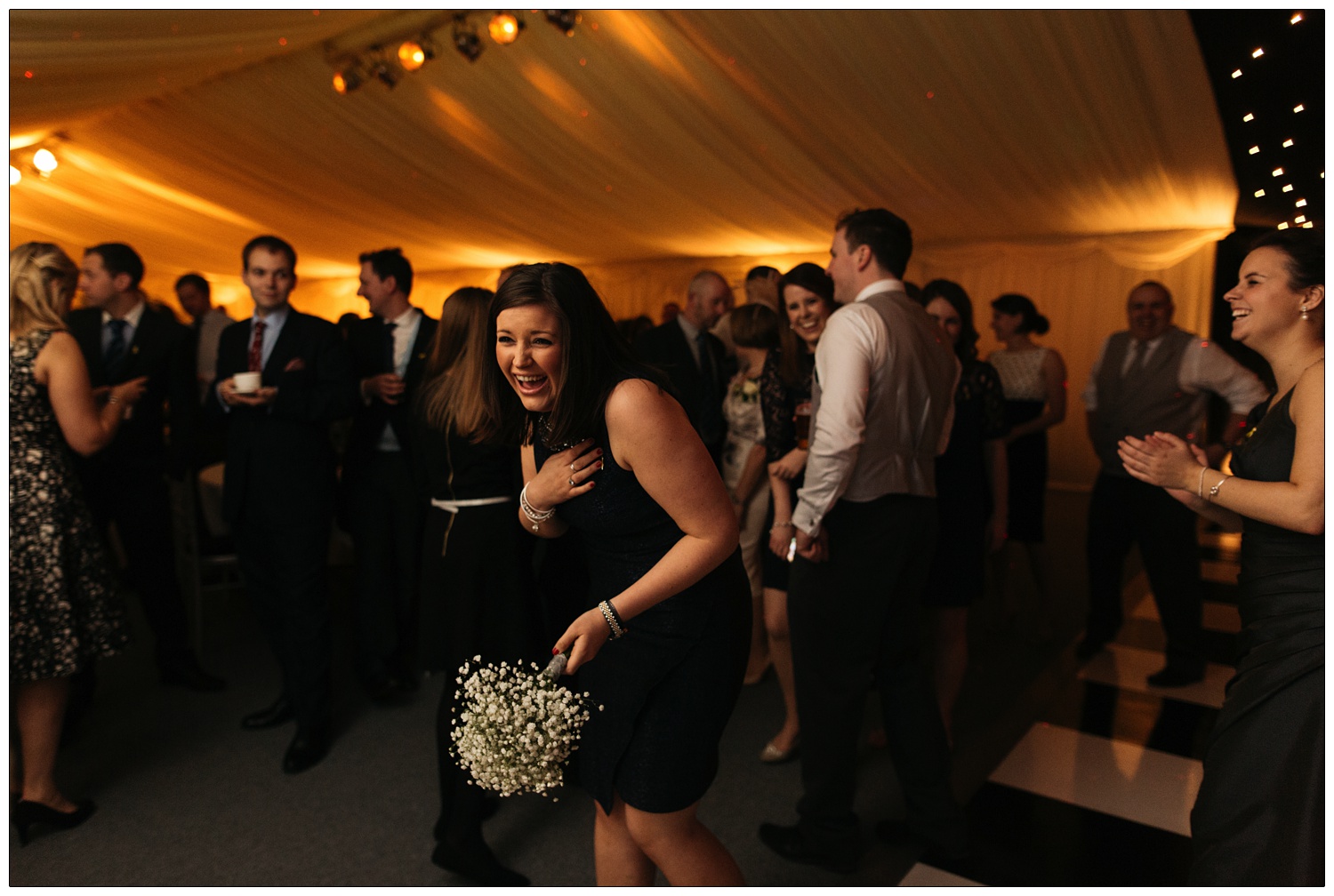 A woman with dark hair in a black dress catches the bouquet and laughs.