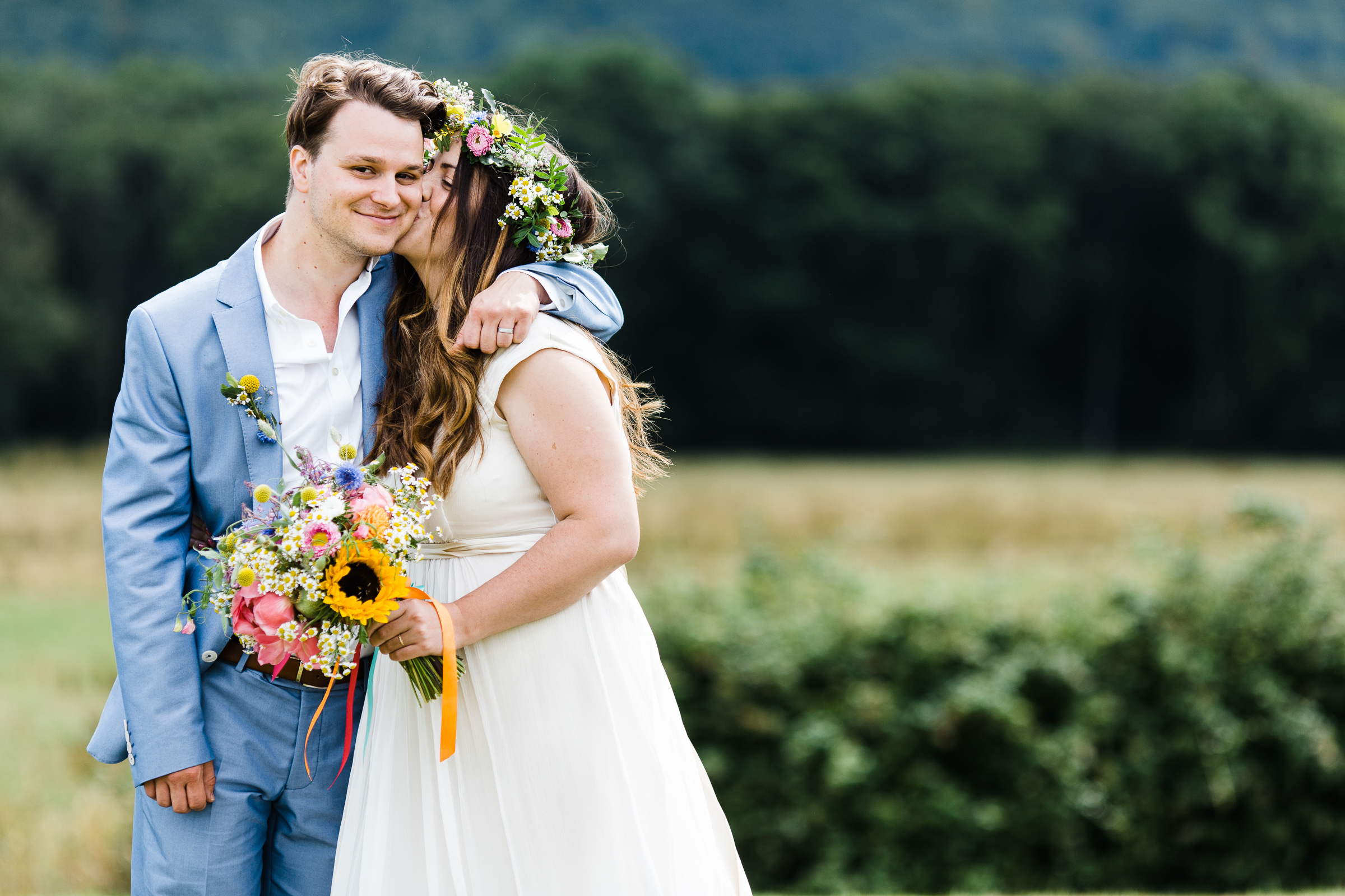 The bride is kissing her new husband on his cheek. She is wearing Saja dress, holding colourful flowers. He is wearing pale blue Zara suit.