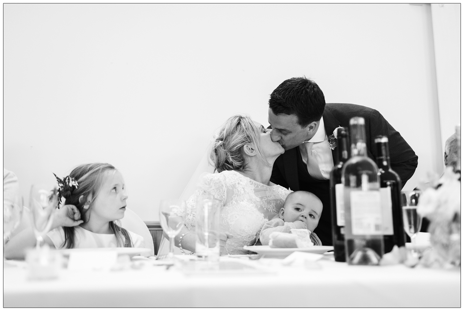 bride and groom kissing while children watch at table.