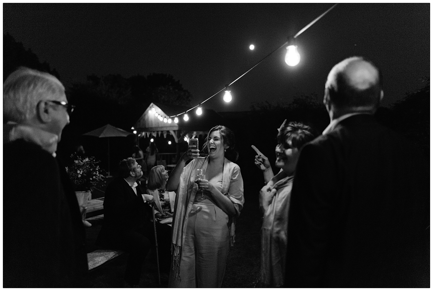 black and white photograph of a laughing woman taking a photo with her iphone under lights outside at night at a wedding reception