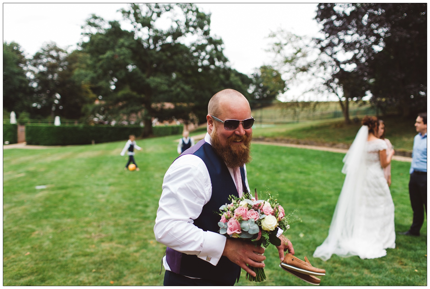 groom holding bouquet outside and new wife and children in the background