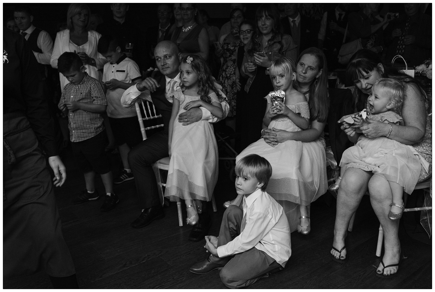 candid picture of people sat on chairs and children sat on their laps, they are watching the first dance and the photography is in black and white