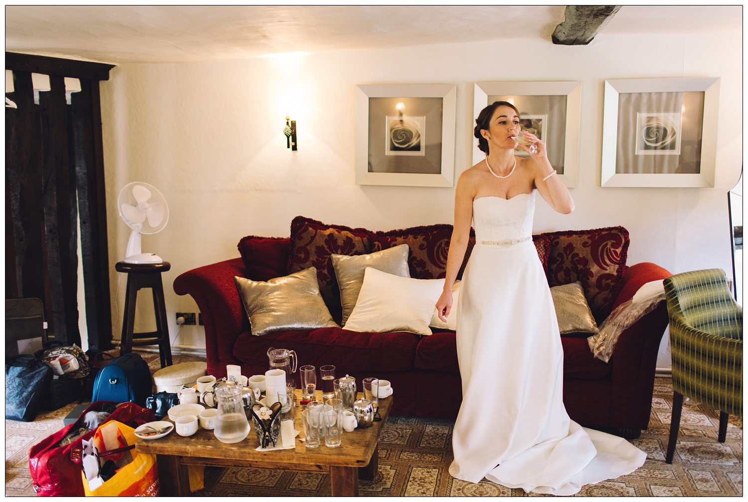 bride having one last sip of prosecco before getting married. She's i her dress, there are glasses and cups on the coffee table. Bags for life on the floor full of items needed to get ready.