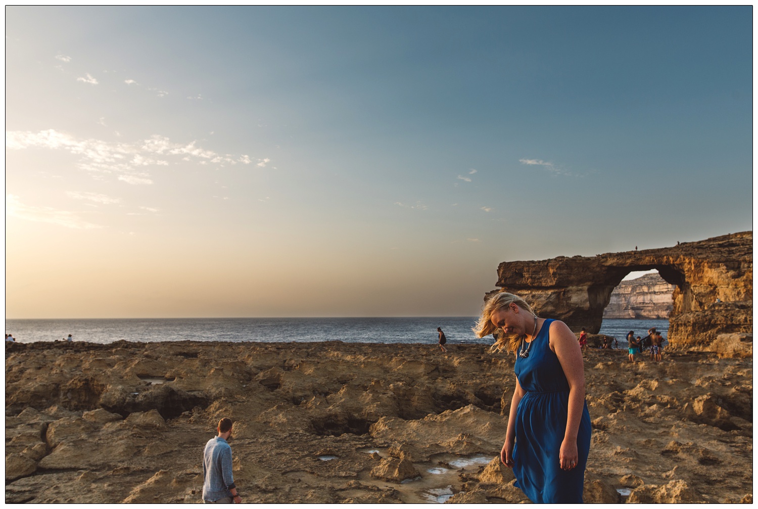 Woman in a blue dress in the foreground with wind in her blonde hair, in front of the Azure Window in Gozo. Her new husband is behind her.