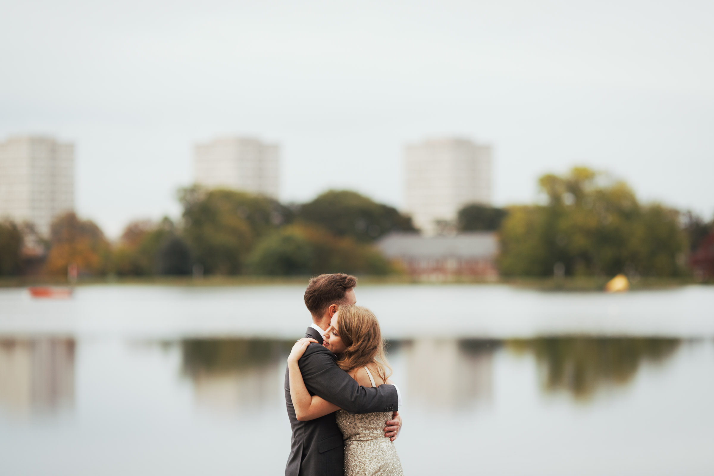Newly married couple hugging by the lake at the West Reservoir Centre in Stoke Newington. the City View Apartments, Devan Grove, N4 in the background. Bride has long hair and is wearing a champagne coloured dress.