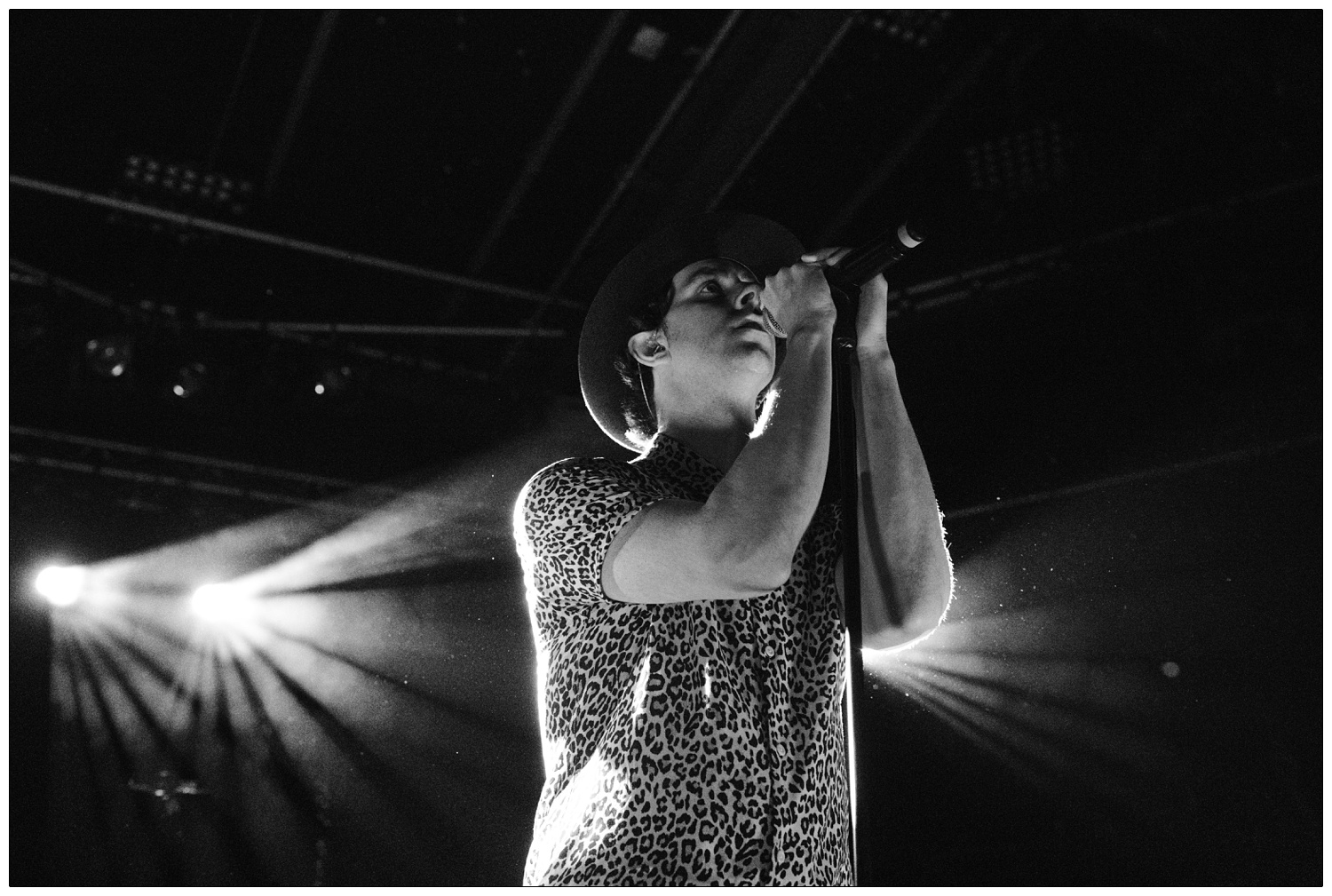 Black and white photograph of Paul Smith holding the microphone at a Maxïmo Park gig. He is wearing a short sleeved leopard print shirt and black hat, a light shines behind him.