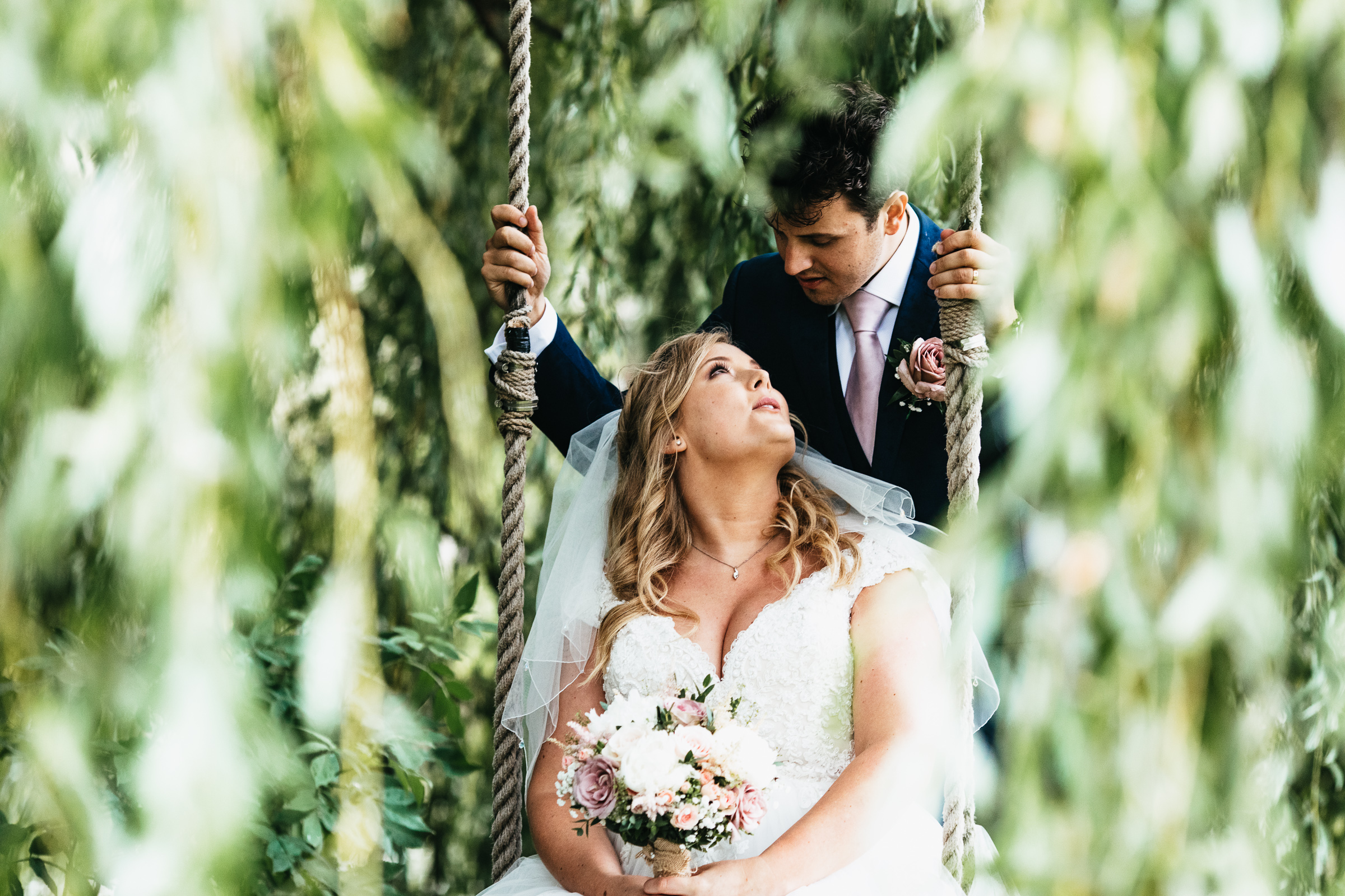 A bride sits on a swing hanging from a willow tree and her groom is behind her. She's looking up at him and holding her flowers. They are in the grounds of Anne of Cleves Barn Great Lodge wedding venue near Braintree.