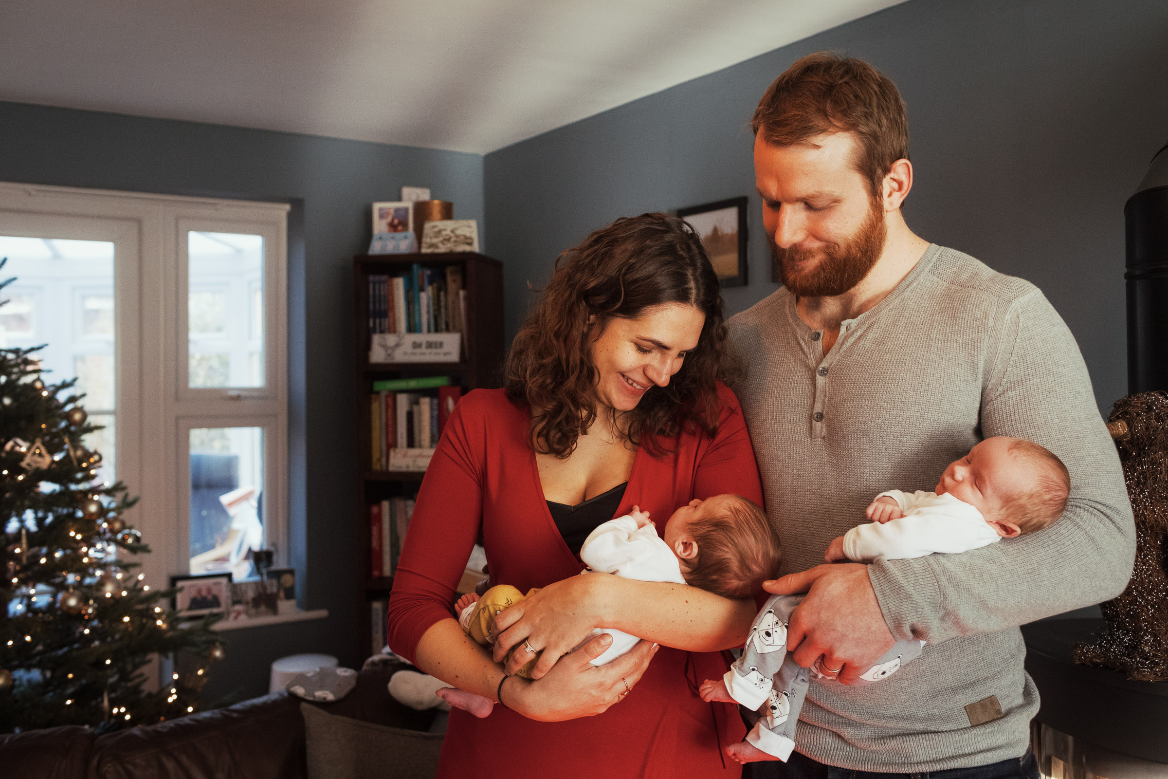 New parents holding their twin babies at Christmas. At home with a christmas tree behind. Family lifestyle photography.