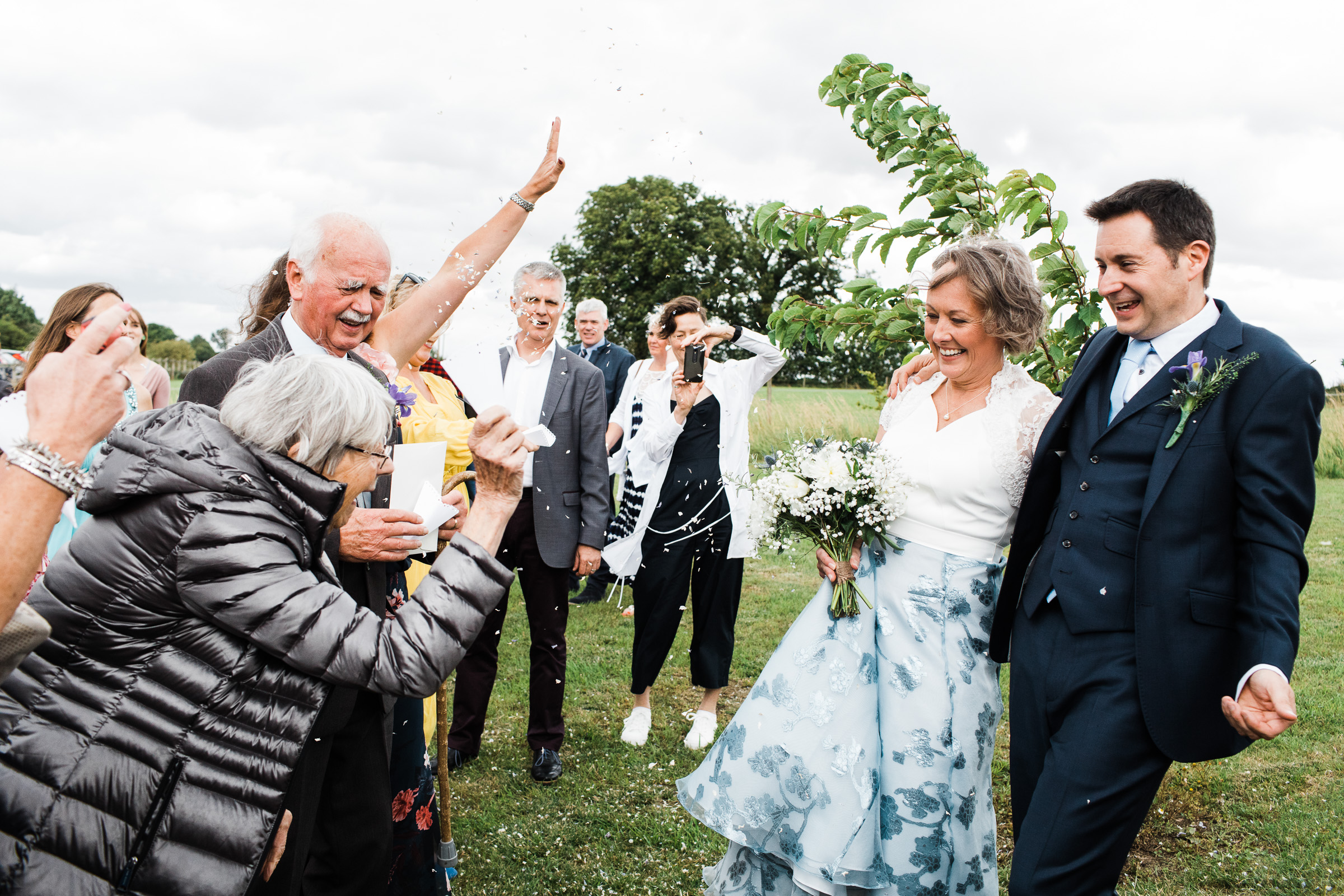 Wedding guests throw confetti at bride and groom. They are outside at Alpheton Hall Barns.