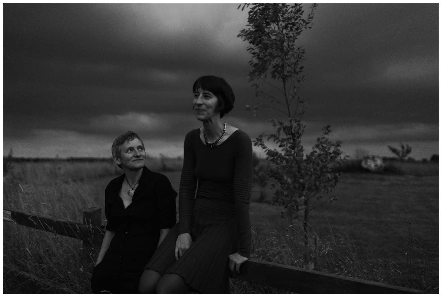 In the dark outside at the Alpheton Hall Barns venue. A woman is sitting on the fence while another woman leans against it and looks up at her.