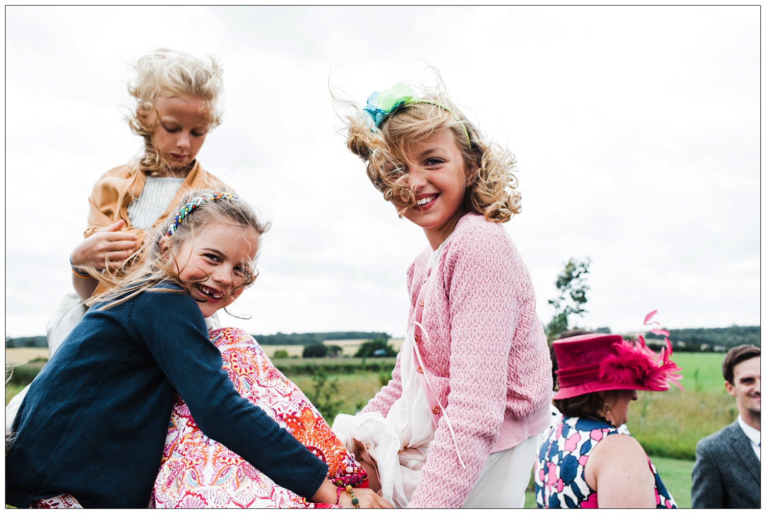 Three girls sat up on a tree trunk in the wildflower arboretum at Alpheton Hall Barns. Two girls are looking at the camera and the wind is blowing their hair around.