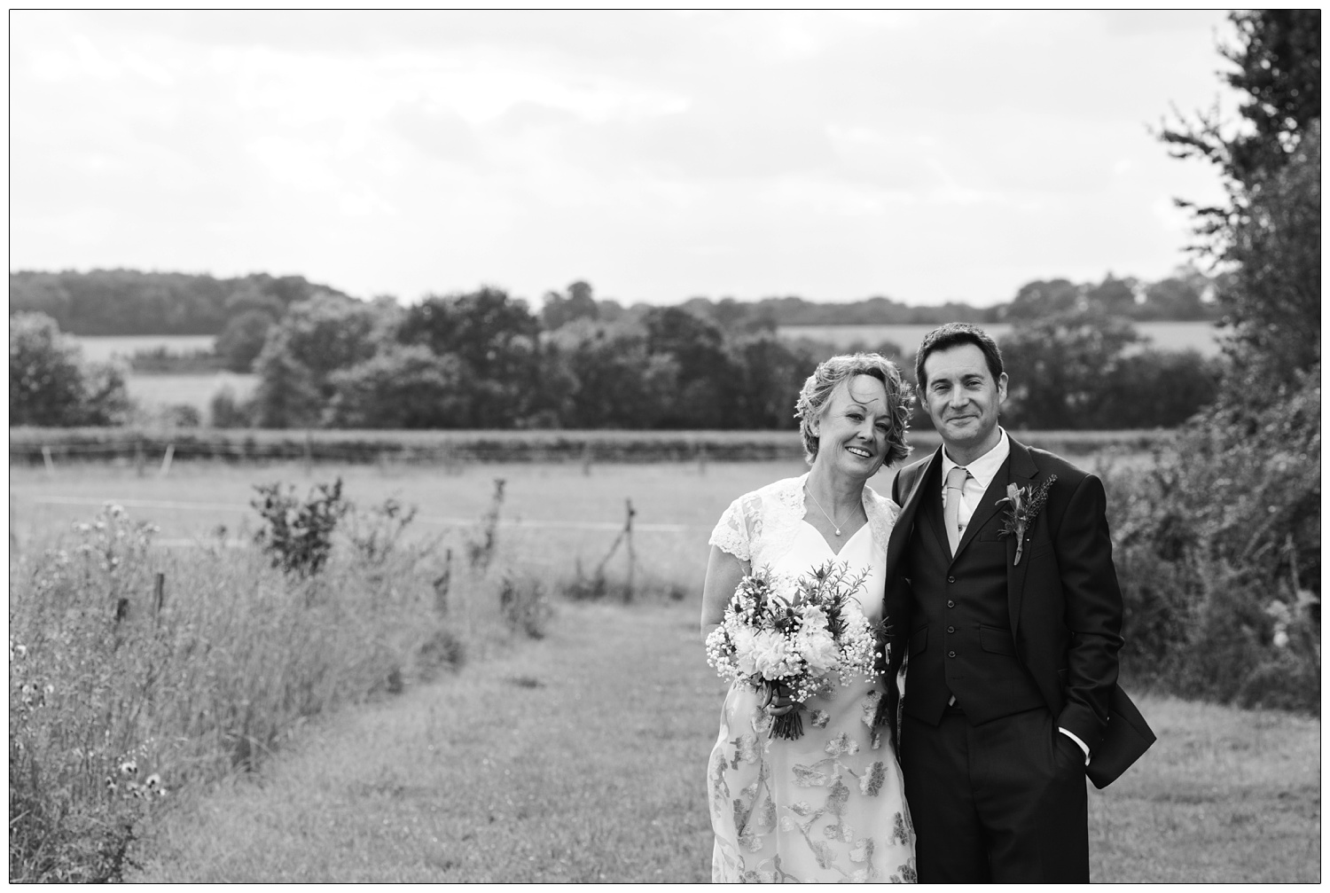 Groom has his hand in his pocket as he stands with his new wife in a grassy lane in the Suffolk countryside.
