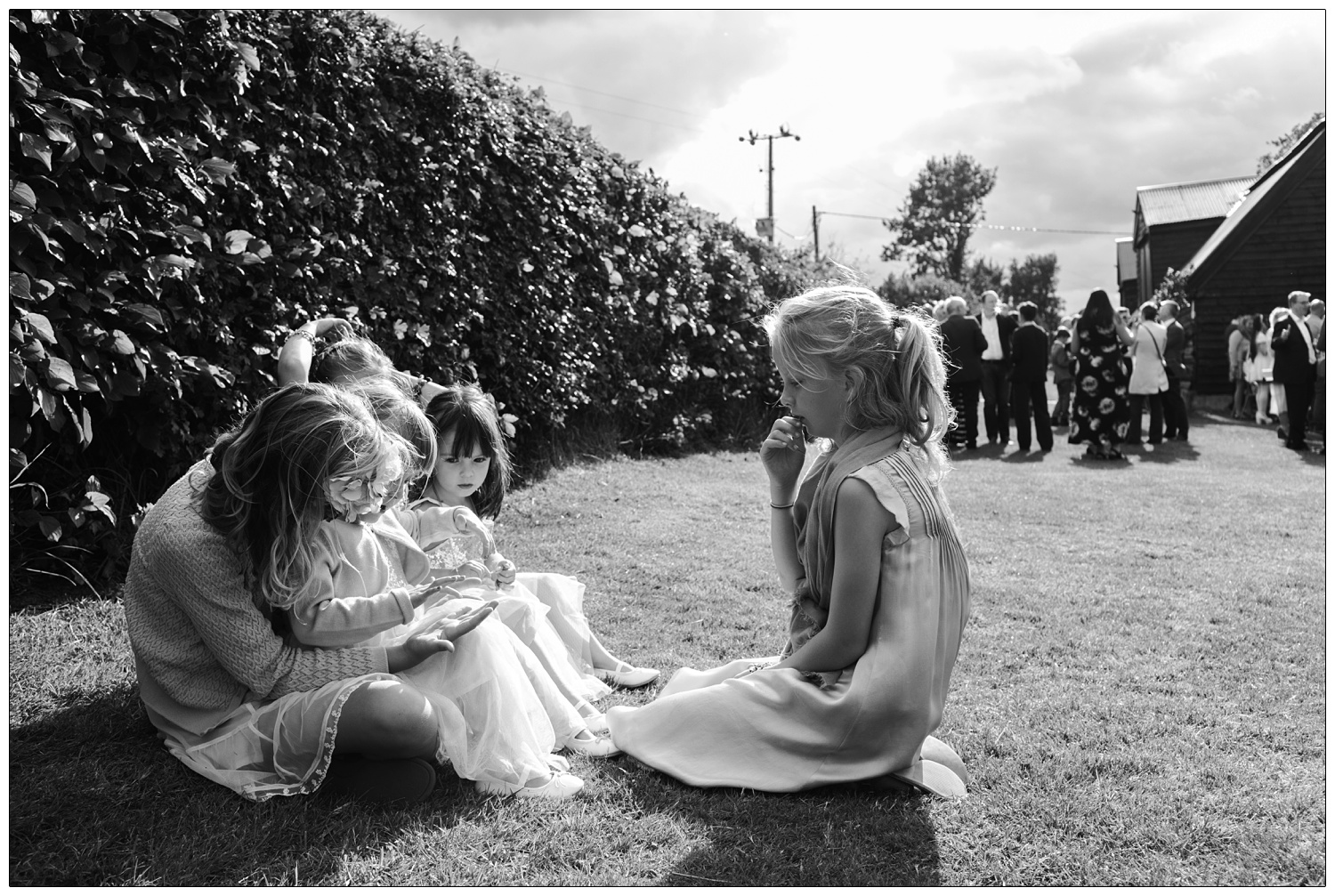 A group of girls are sat in the wedding garden by a hedge. Adults are in the background. they are at Alpheton Hall Barns.