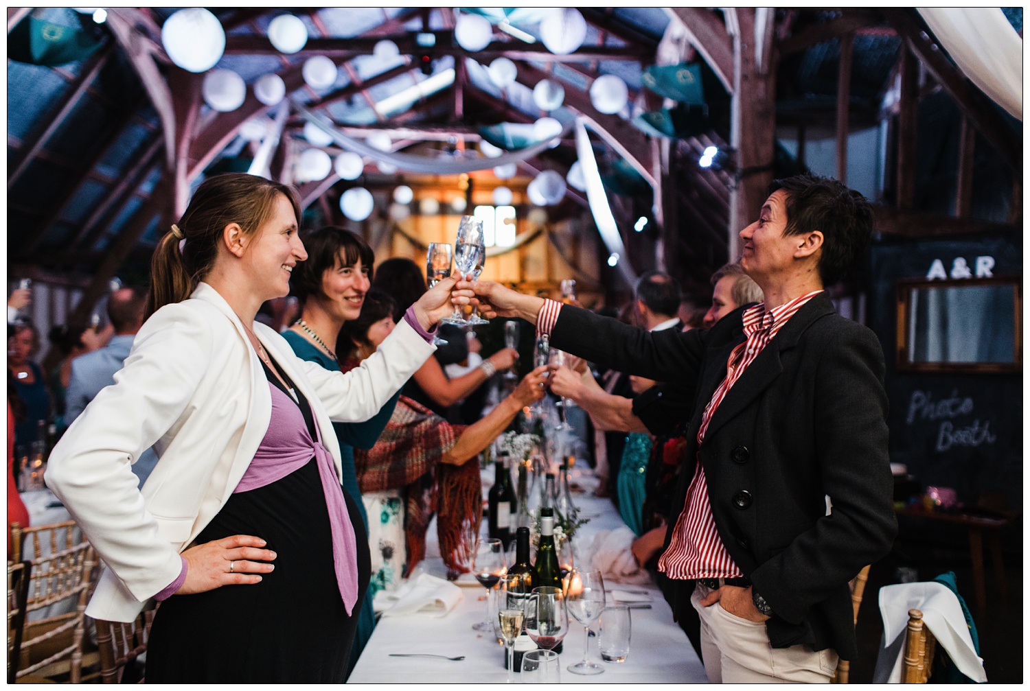 Wedding guests standing at a long table toasting with their drinks. The ceiling of Alpheton Hall Barns is decorated with round paper lanterns and lighting.