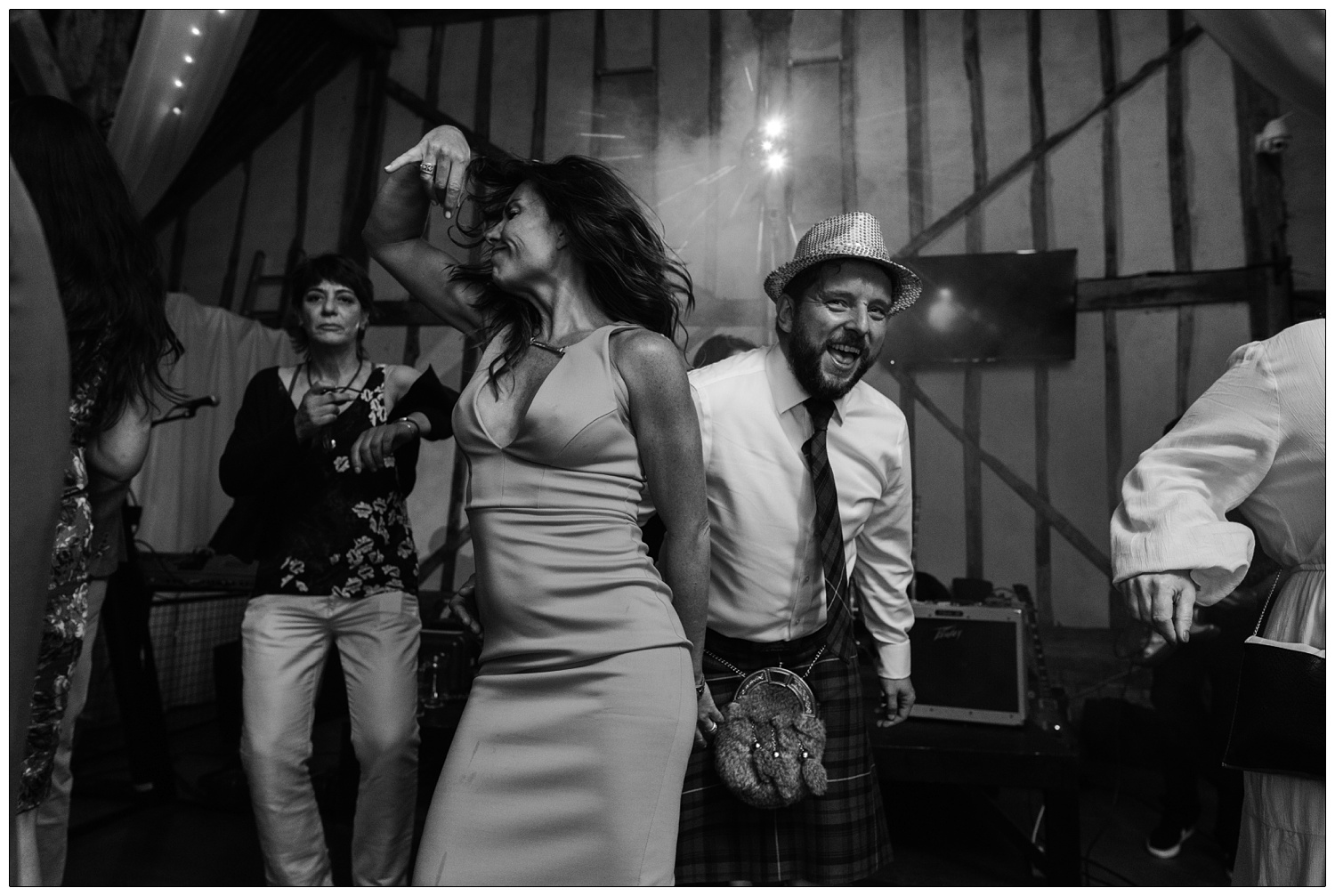 A man in a kilt and sparkly hat is dancing behind a woman in a dress and there are lights shining behind them. A candid style photograph at a wedding reception.